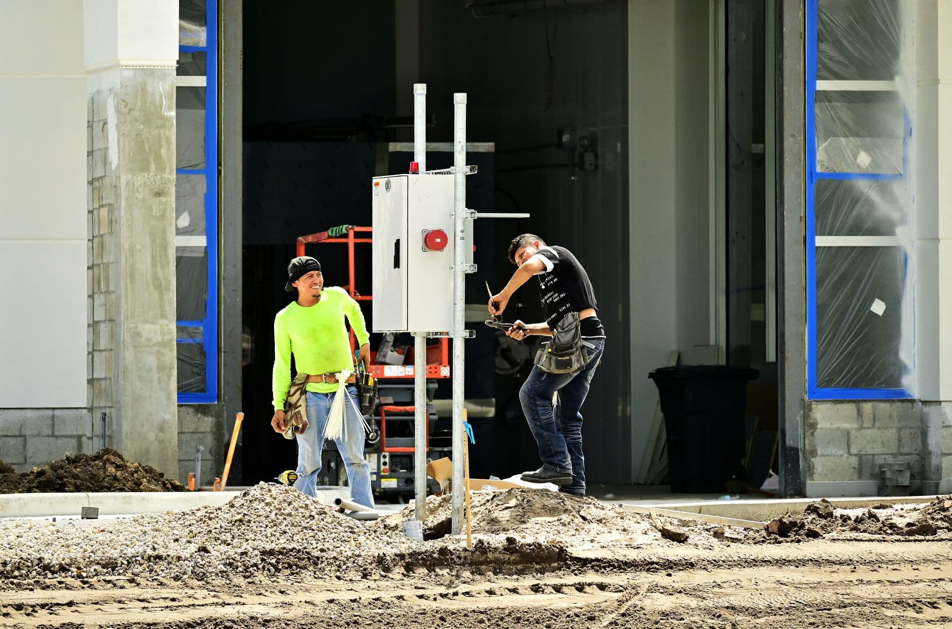 Two workers wiring a temporary power panel on a post outside a building under construction