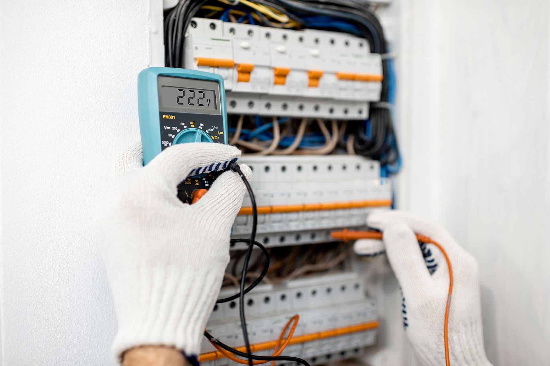 Gloved hands holding a digital multimeter against the breakers inside an open electrical panel
