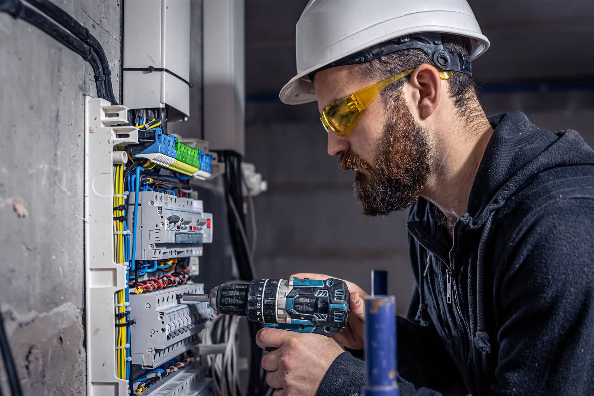 An electrician in a hard hat and safety glasses driving a screw into a breaker inside an open switchboard