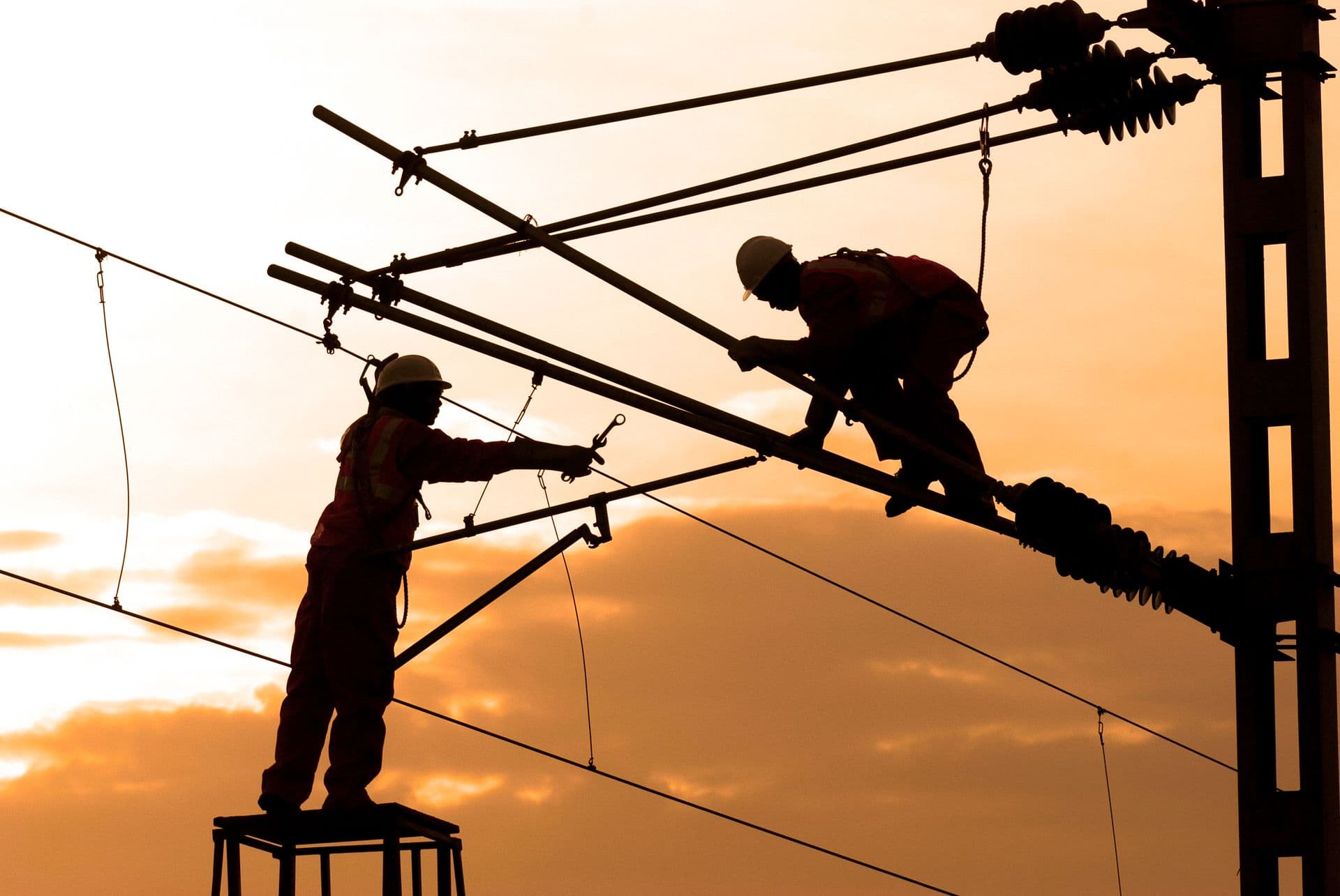 Two electricians silhouetted against an orange sky while working on overhead lines