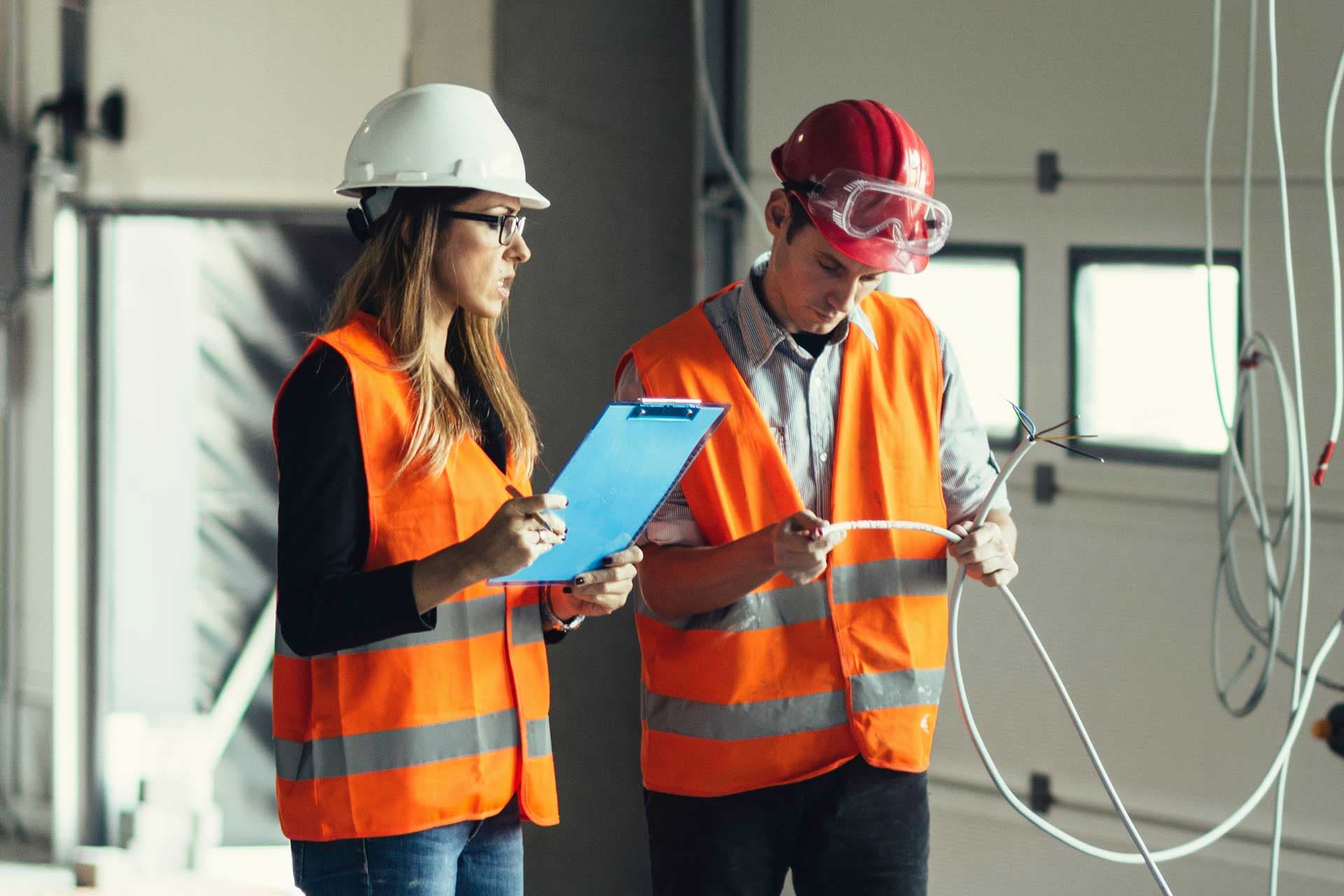 Two workers in high-visibility vests checking a cable against notes on a clipboard in an unfinished building