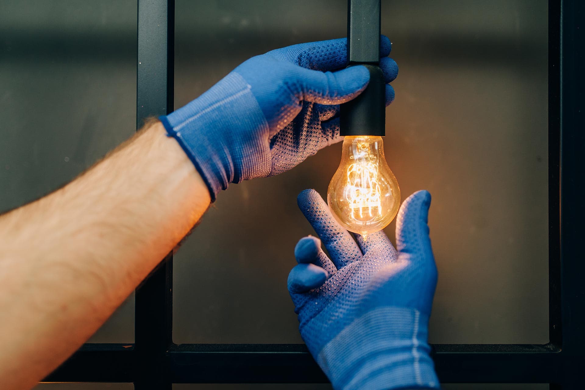 Gloved hands fitting a lit filament bulb into a black pendant lampholder