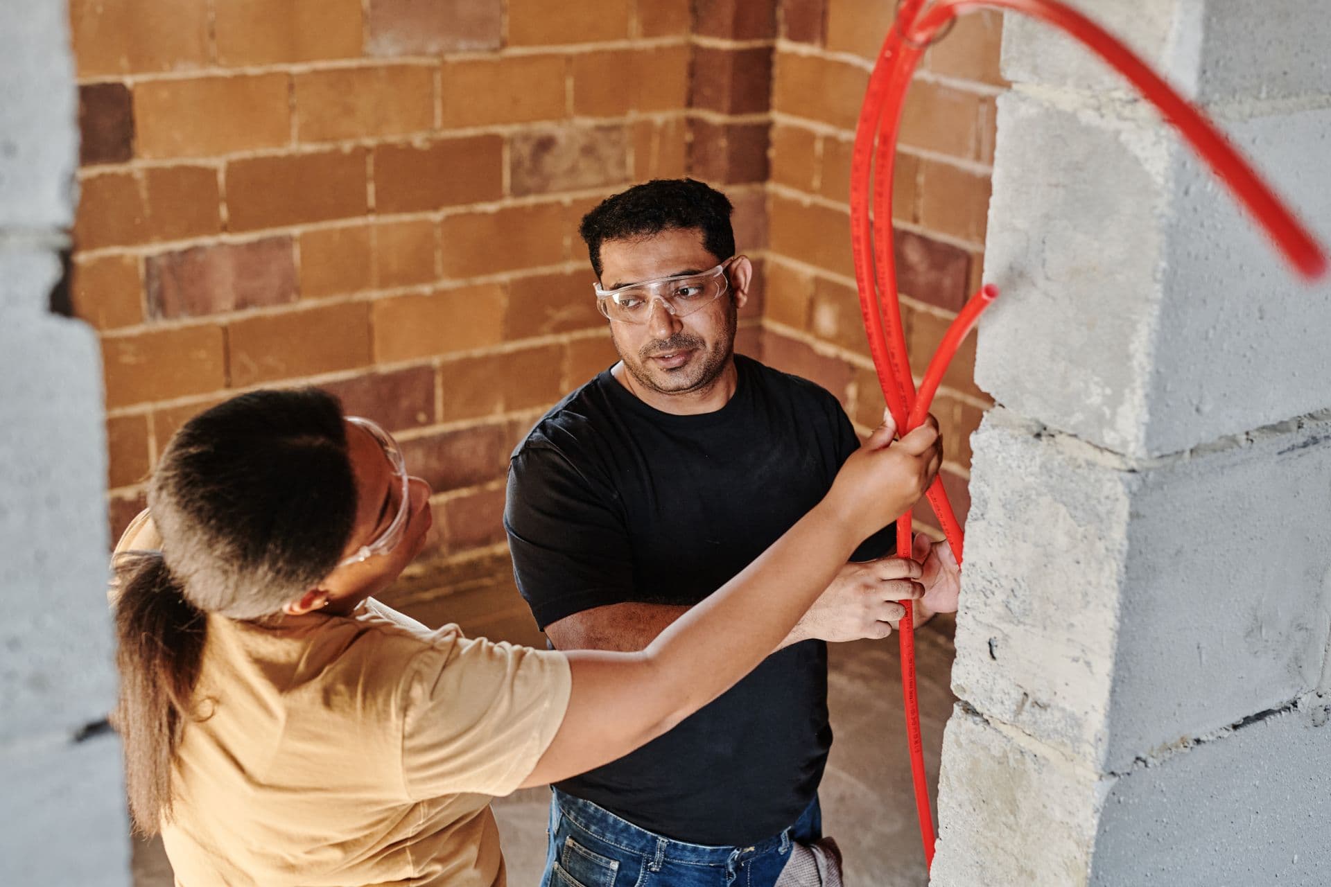 Two electricians routing new red conduit and cable through a block wall