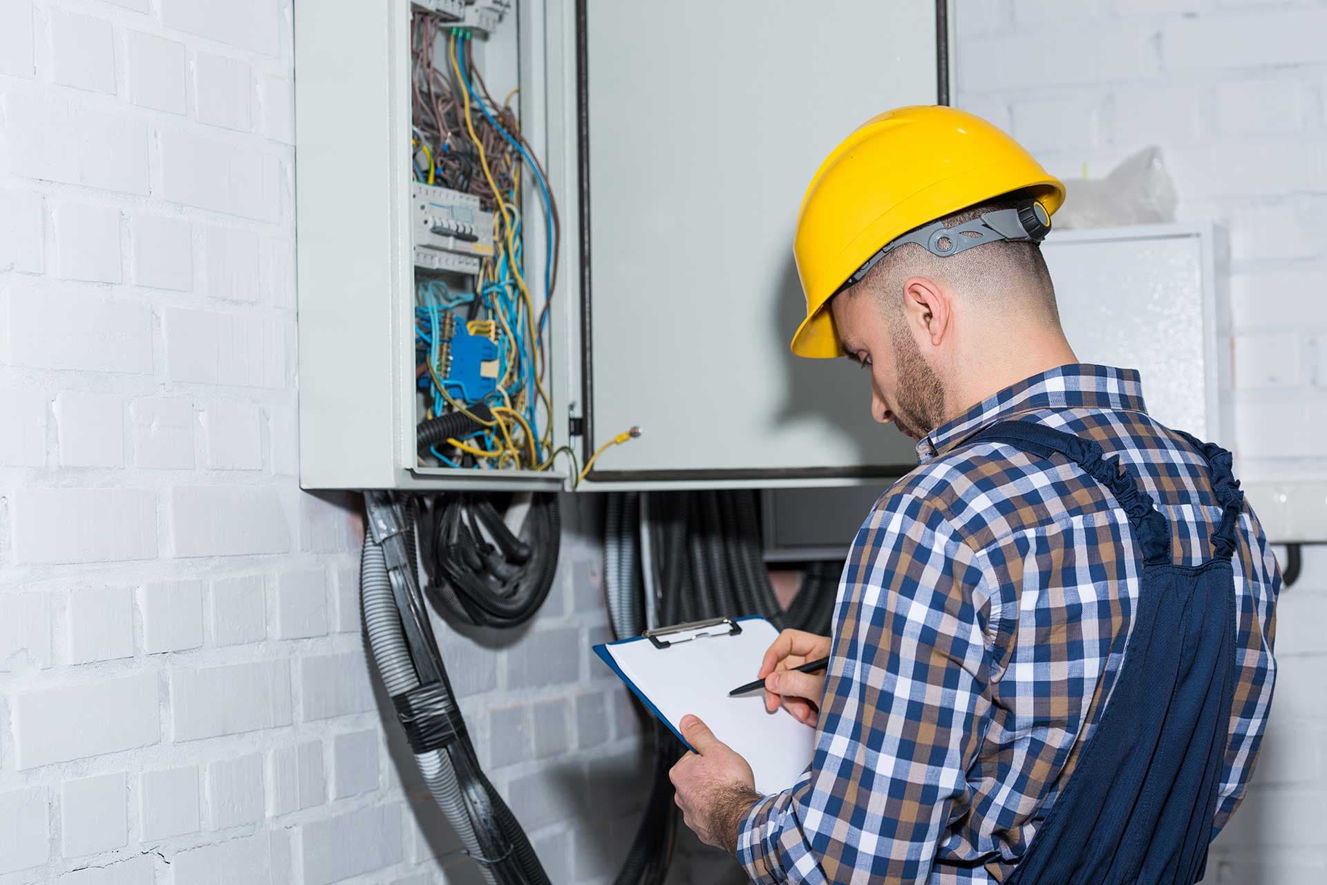 An electrician writing on a clipboard beside an open wall enclosure packed with wiring and conduit