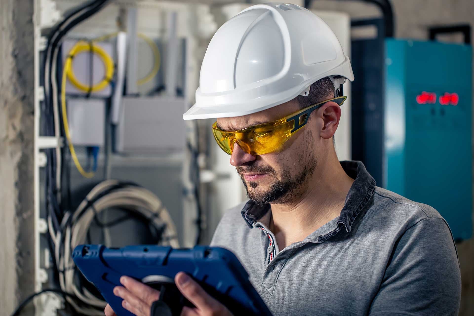 A technician in a hard hat reading a tablet beside control panels in a plant room